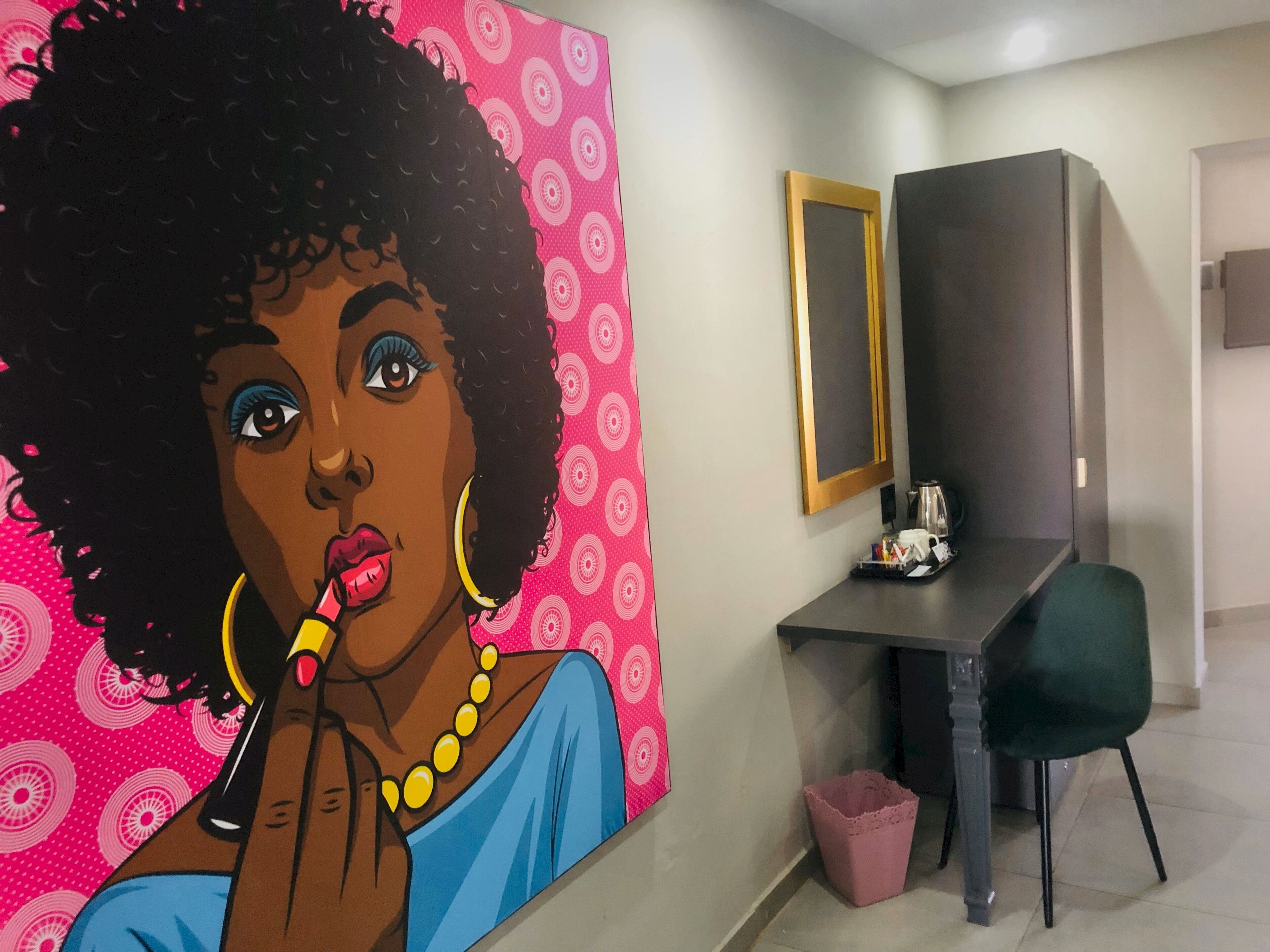 Une chambre d’hôtel avec un grand tableau au mur, un bureau avec une chaise, une poubelle et un miroir.