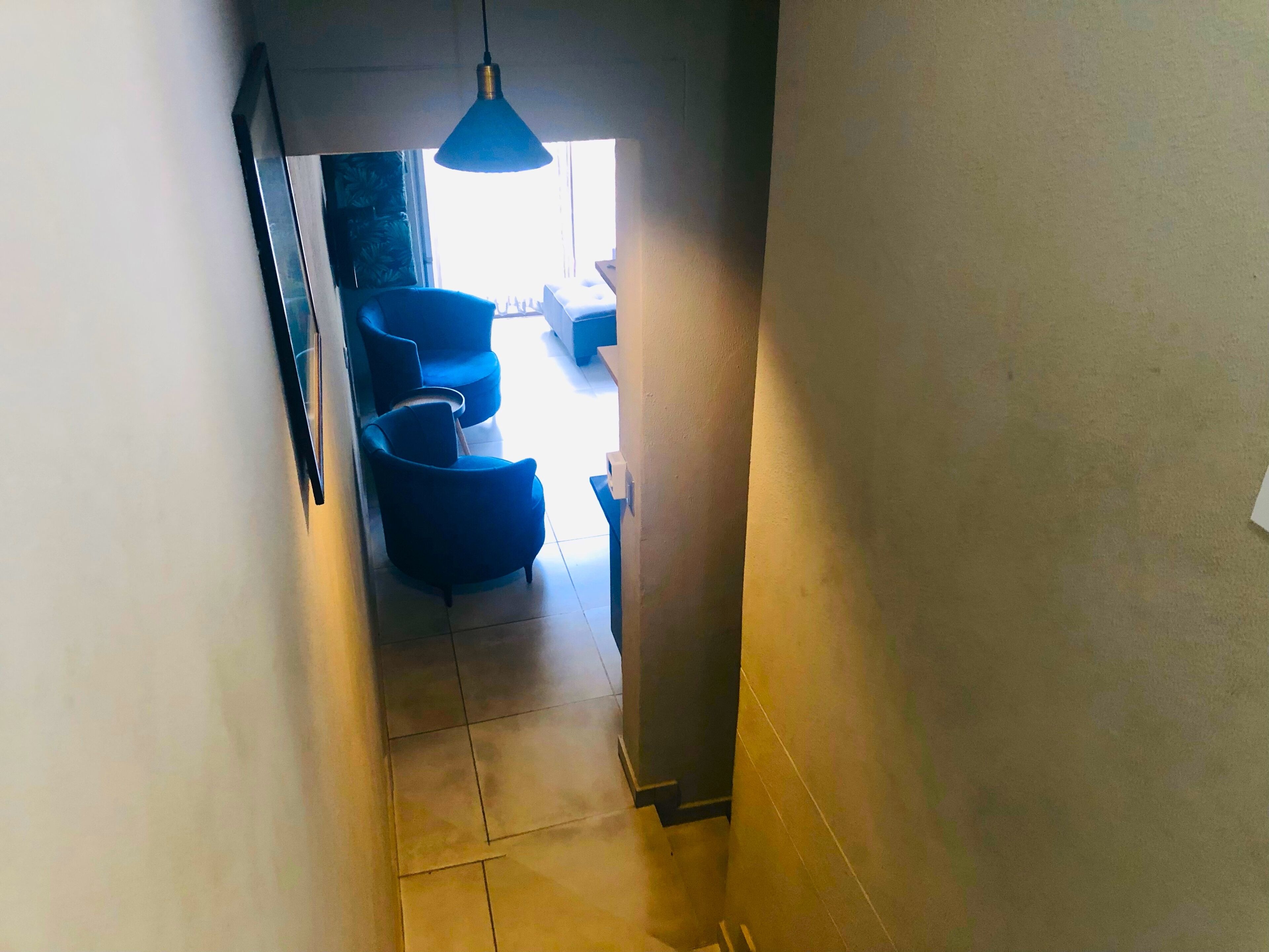 Um corredor estreito com piso ladrilhado que leva a uma área de estar com cadeiras azuis e um lustre.