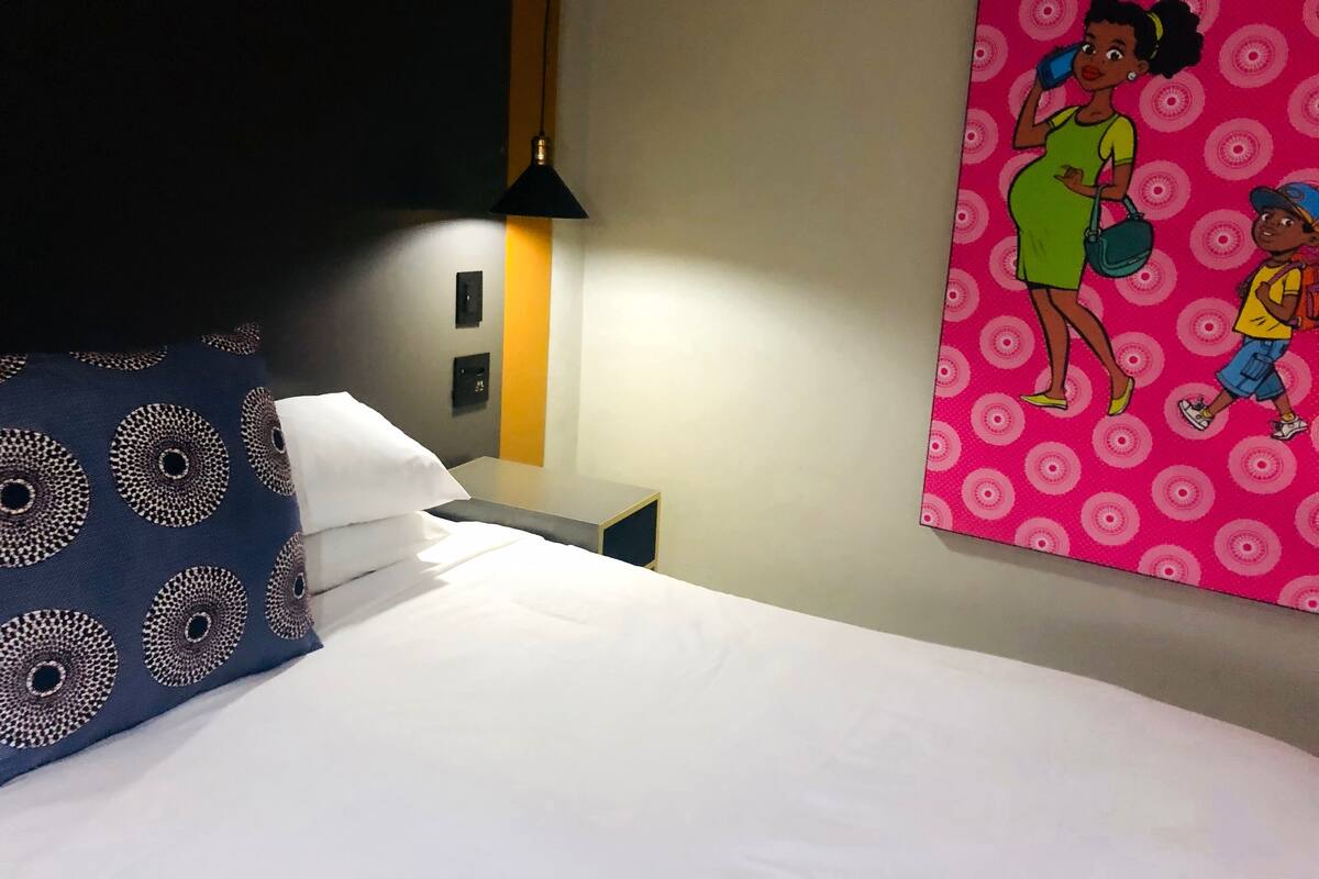 Une chambre d’hôtel avec un lit, une table de chevet, une lampe et un mur orné d’une illustration colorée.