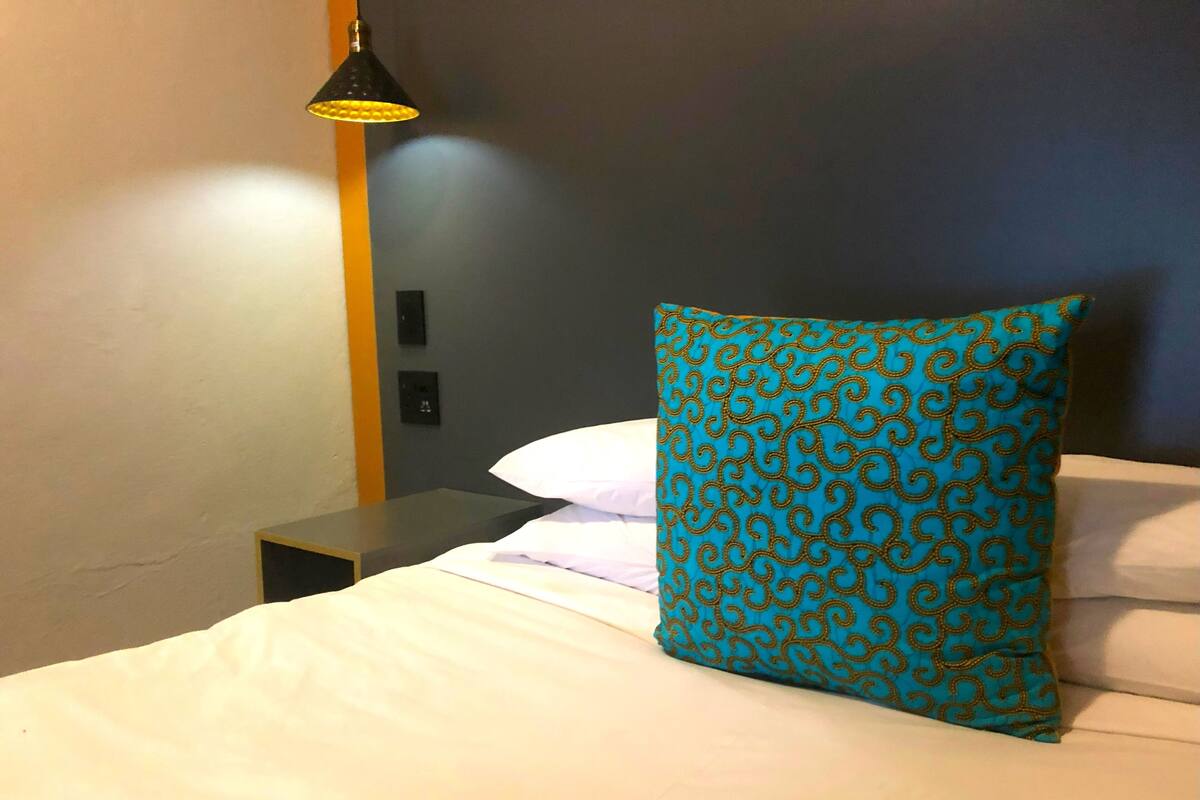 Un lit bien fait, avec du linge de lit blanc et un seul coussin à motifs turquoise. Une table de chevet noire, sur laquelle se trouve une lampe noire, est placée à côté du lit.