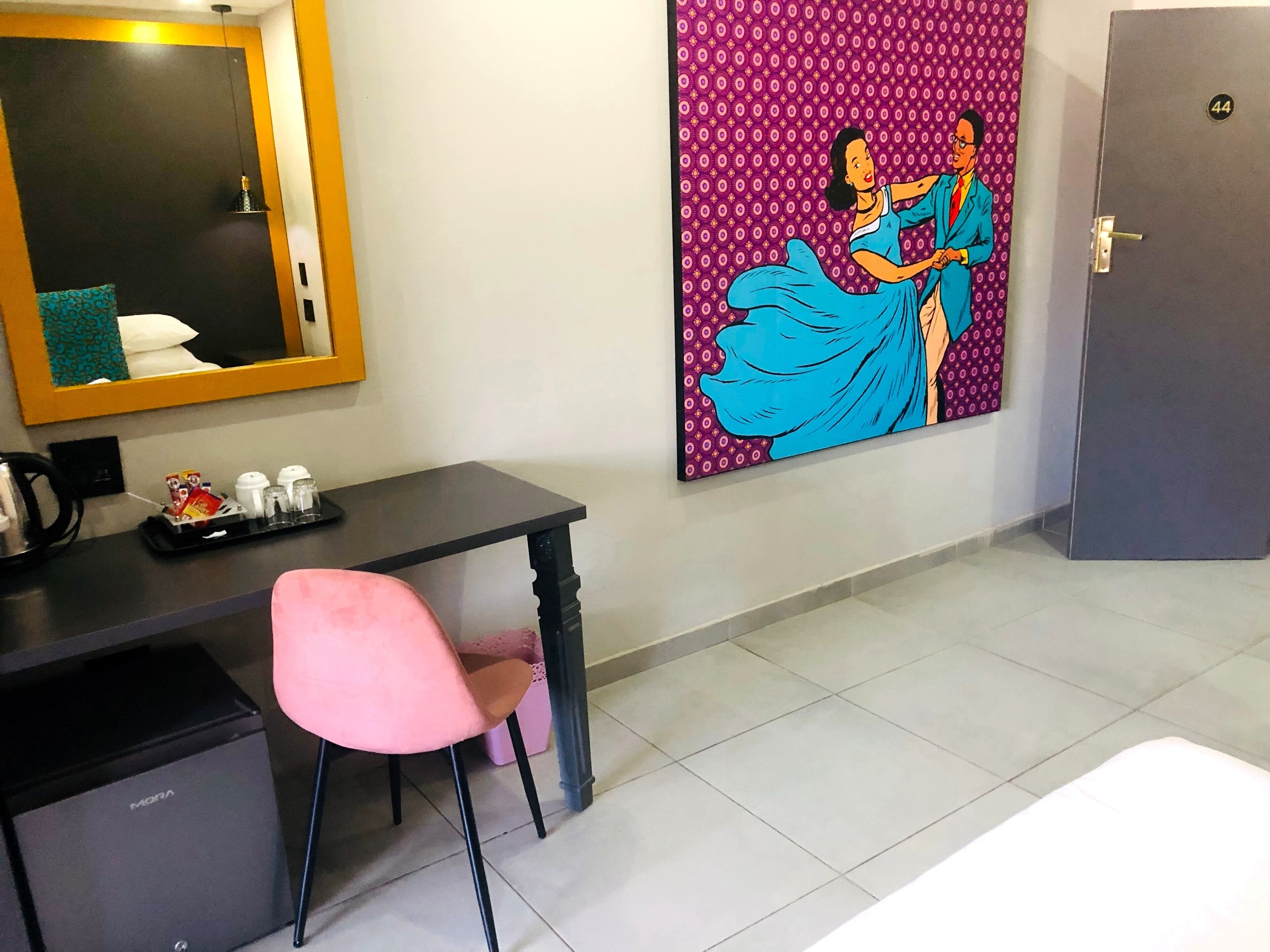 Una habitación de hotel con escritorio, una silla rosa, un mini bar, un espejo y una obra de arte colorida en la pared.