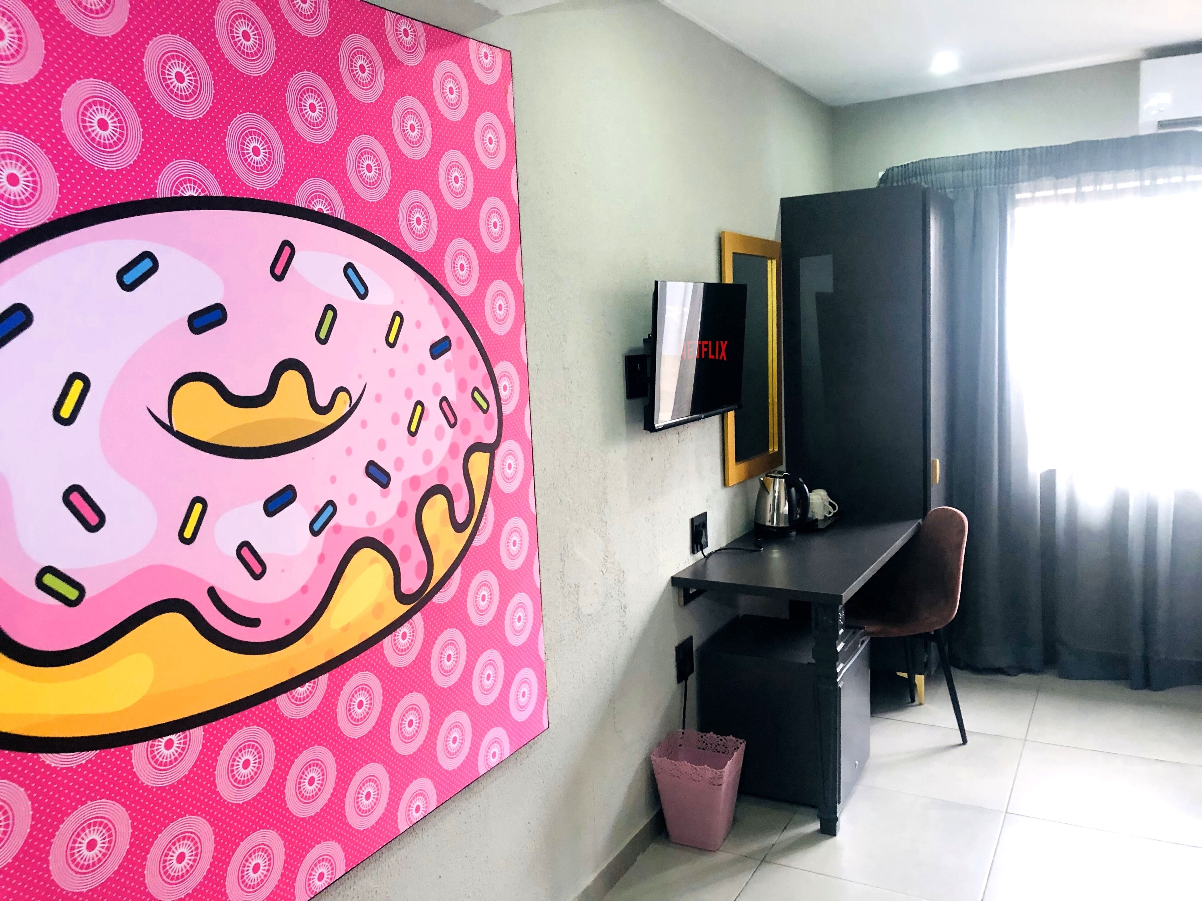 A room with a large pink wall mural, a flat-screen TV, a desk with a chair, a coffee maker, and a trash bin.