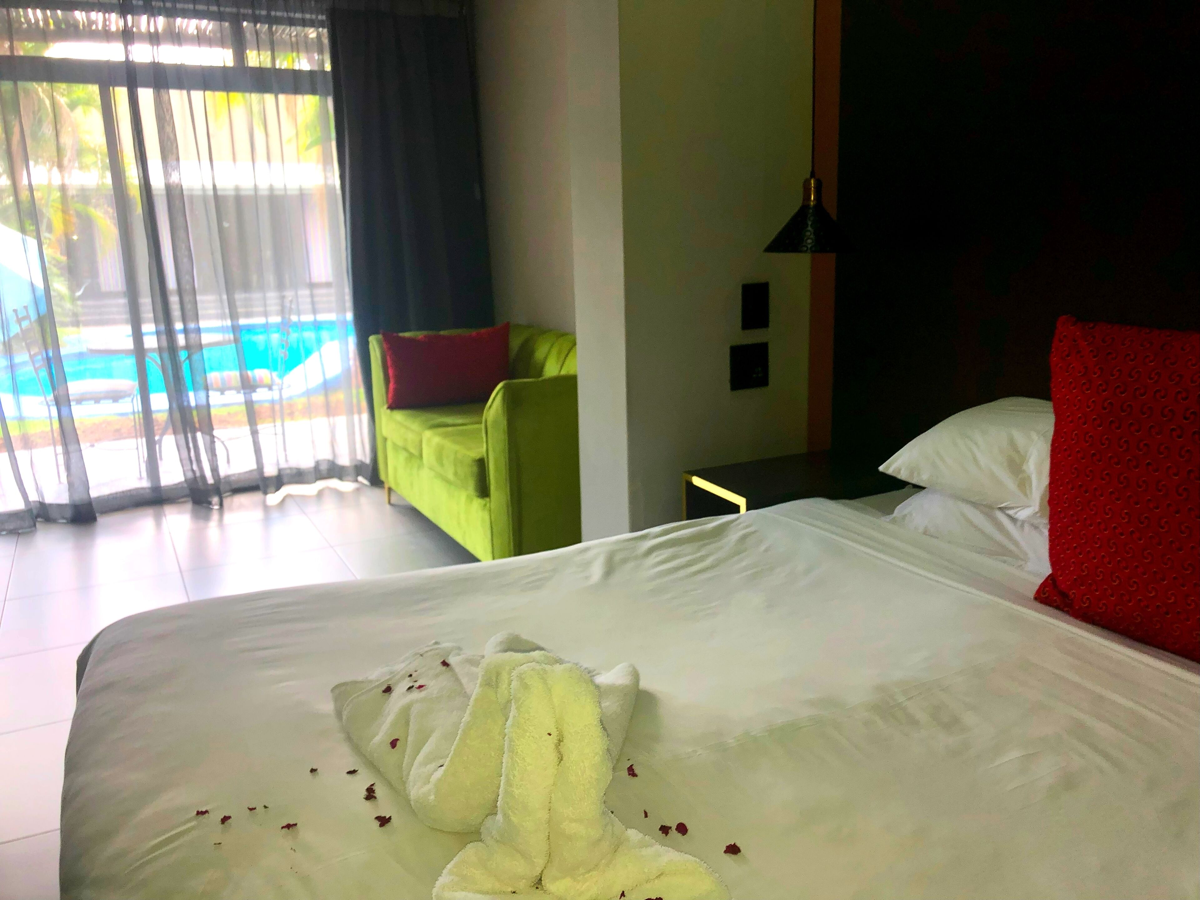 Habitación de hotel con cama, un sillón verde, un cojín rojo y vista a una piscina a través de cortinas translúcidas.