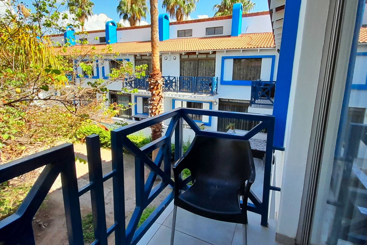 Un balcon avec une chaise noire, une vue sur un immeuble aux accents bleus et des palmiers en arrière-plan.