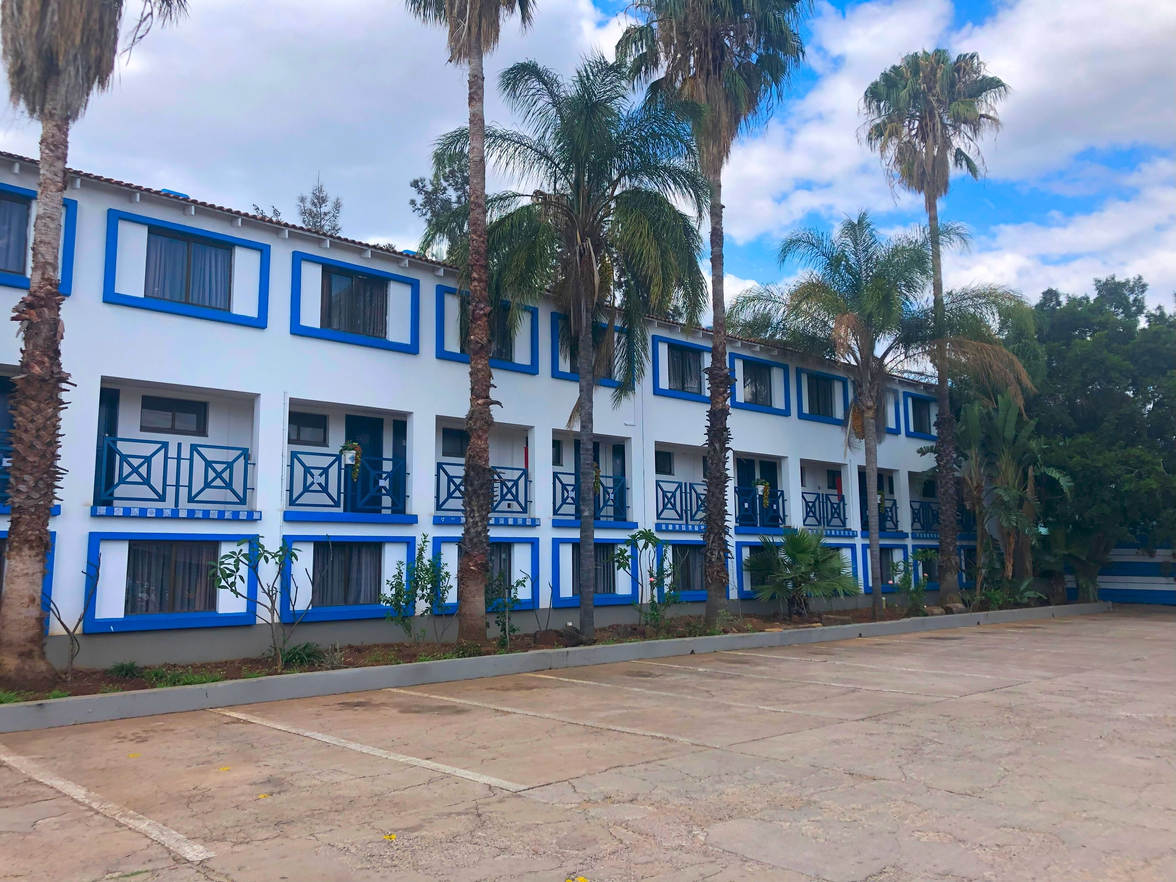 A multi-story hotel with blue-framed windows and balconies, surrounded by palm trees.