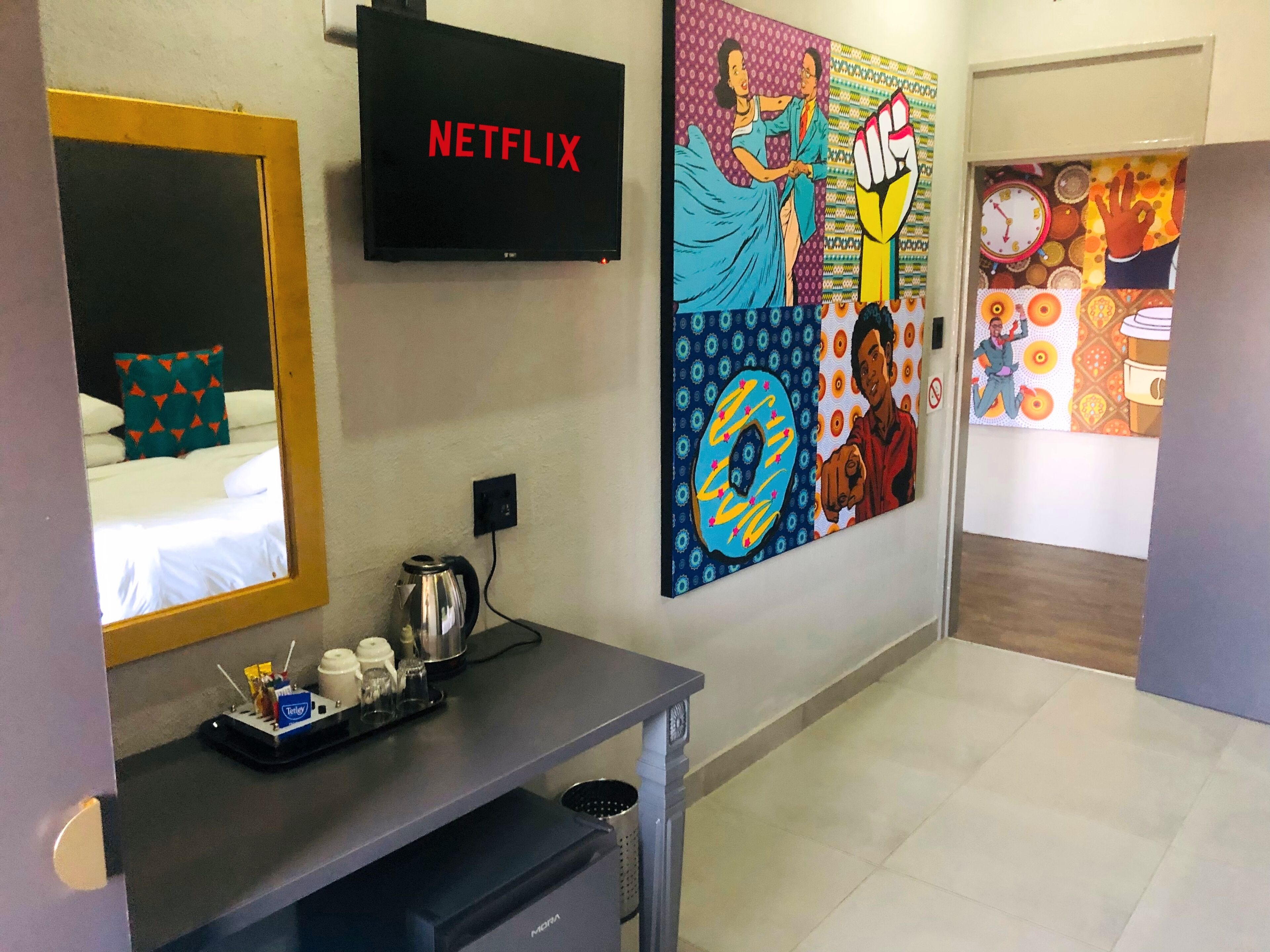 A hotel lobby with a TV displaying 'NETFLIX', a coffee machine, a tray with condiments, and a colorful mural on the wall.