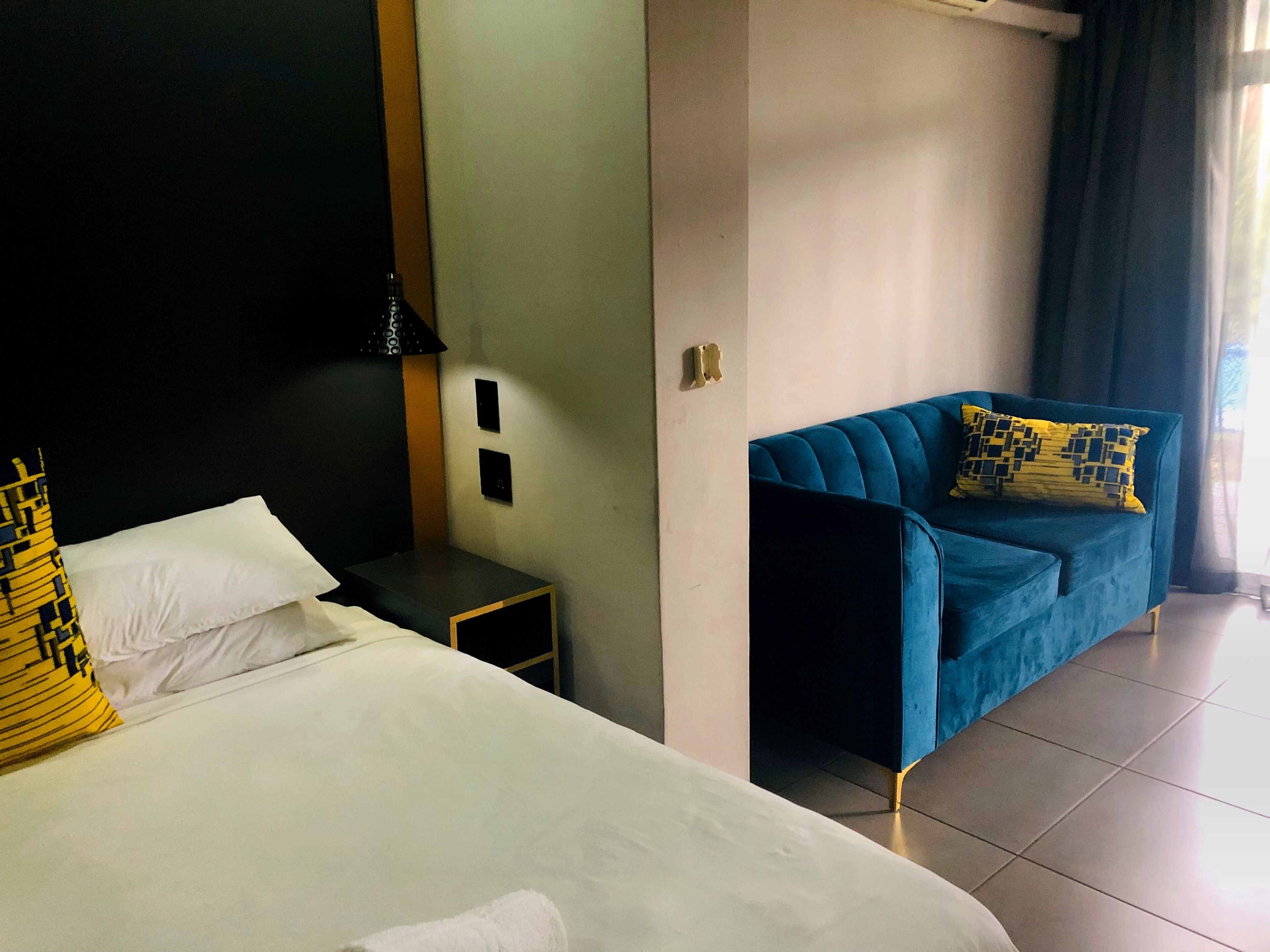 A hotel room with a bed, a nightstand, a blue sofa, and a window with curtains.