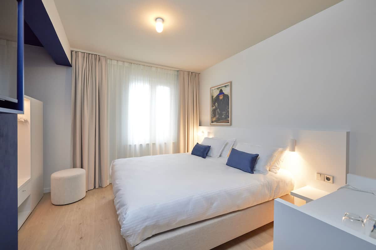 Una habitación de hotel moderna con una cama grande, mesitas de noche, un otoman pequeño y redondo, y un cuadro en la pared.