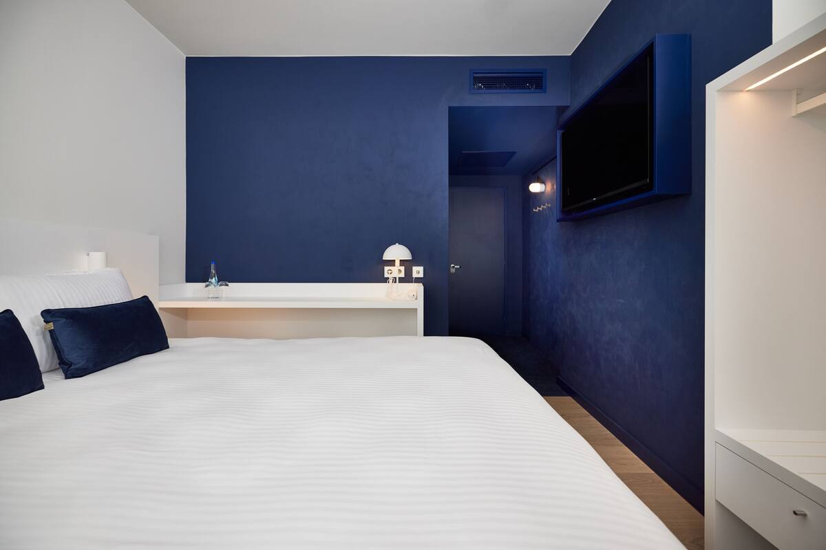 Habitación de hotel moderna con una cama grande, una mesita de noche con lámpara, un televisor montado en la pared y una pared de acento azul oscuro.