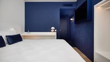 A modern hotel room with a large bed, a bedside table with a lamp, a TV mounted on the wall, and a dark blue accent wall.