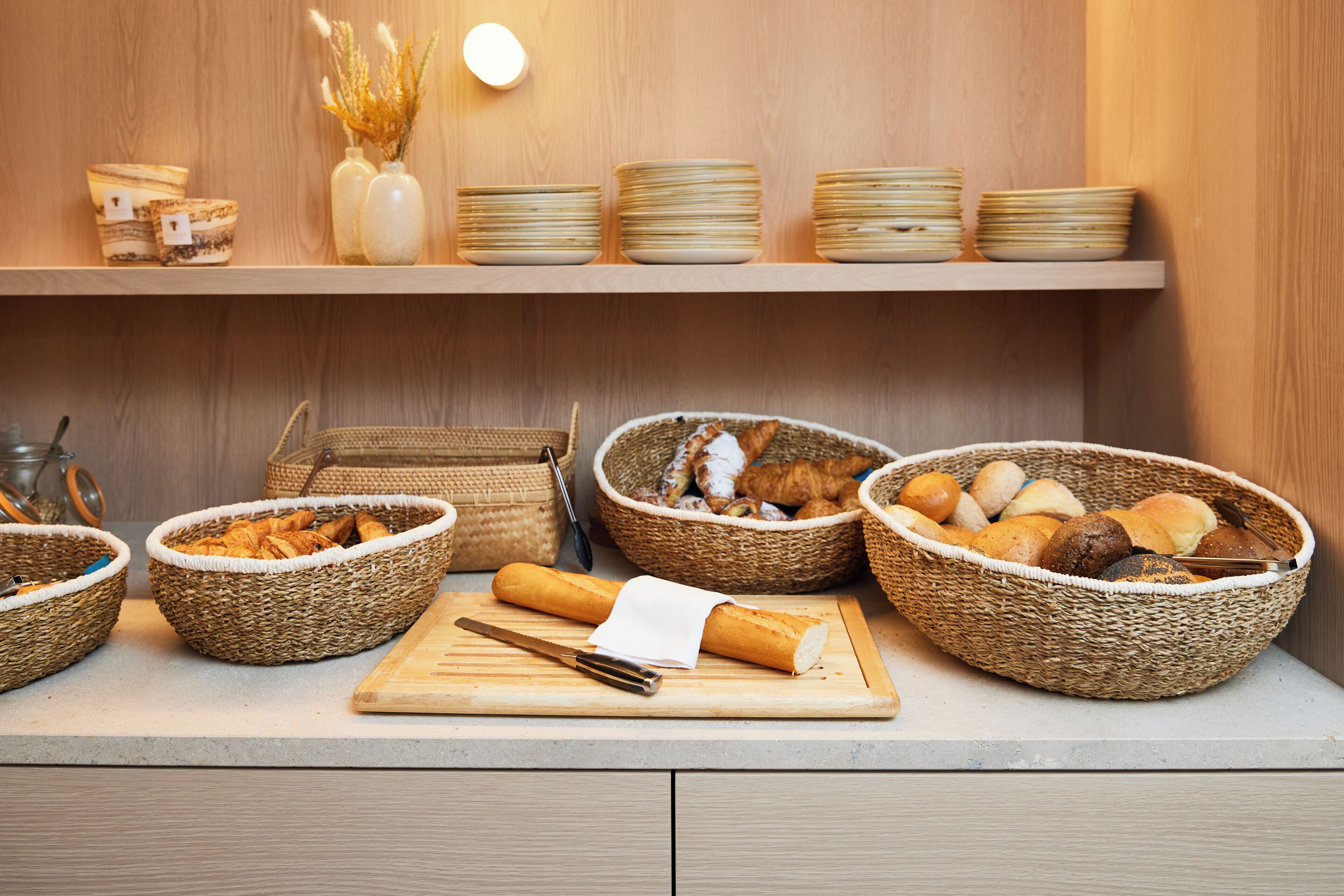 Een ontbijtbuffet met broden en gebak, een snijplank met mes, en gestapelde borden op een houten plank.