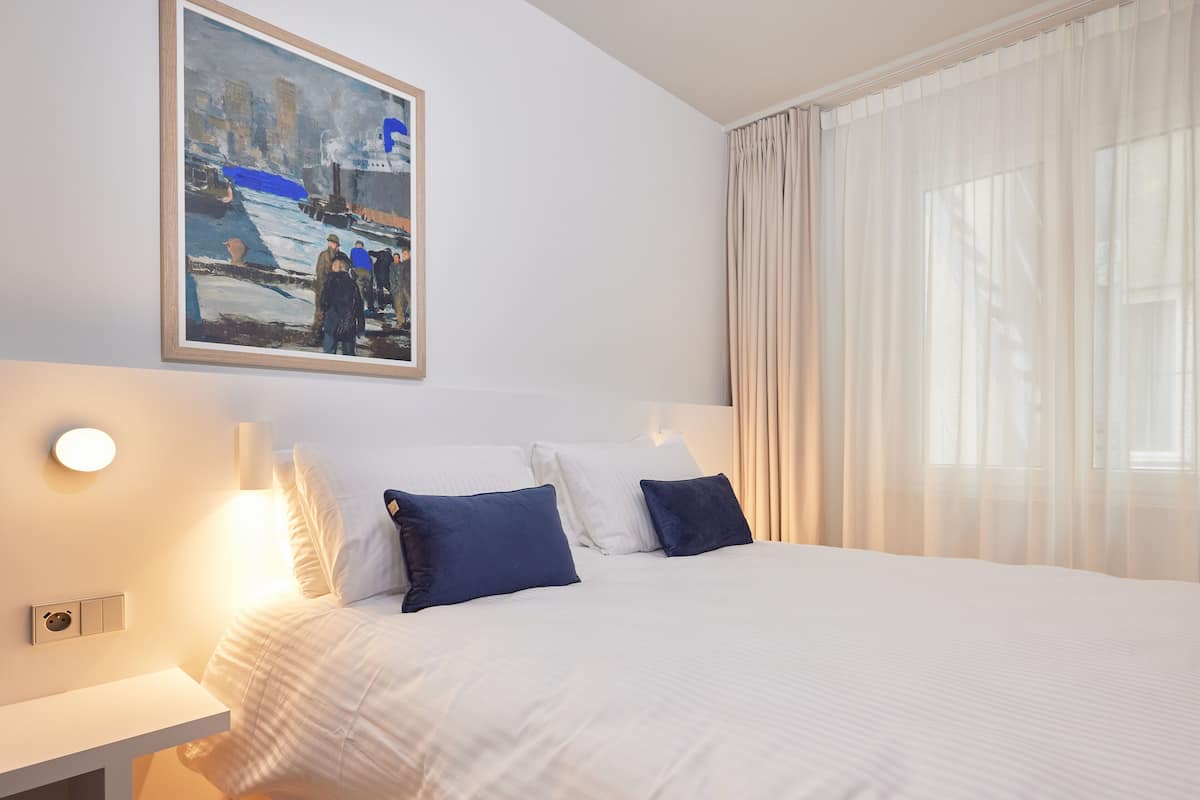 Habitación de hotel con cama, ropa de cama blanca, un cuadro en la pared y una ventana con cortinas de gasa.