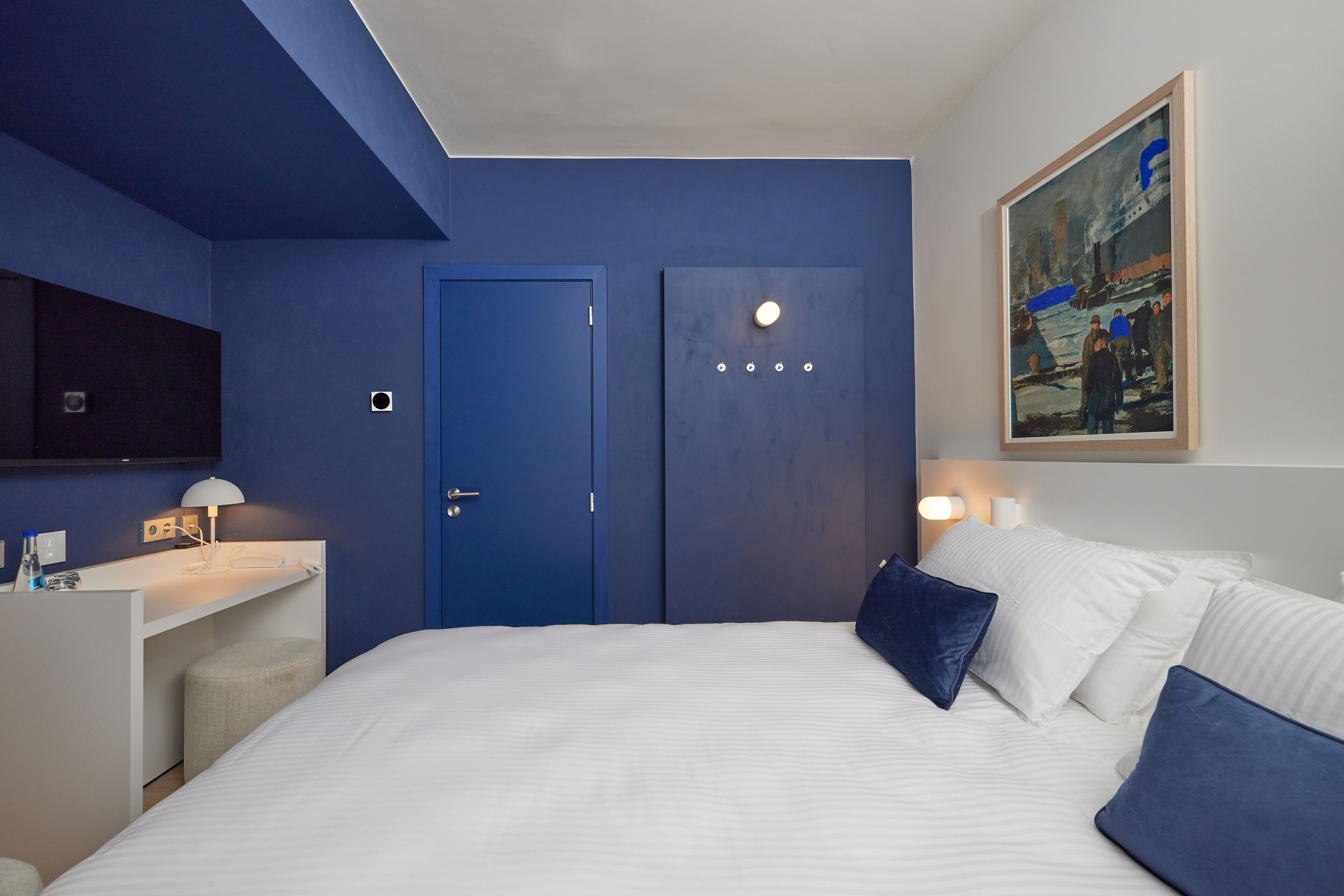 Habitación de hotel con una cama grande, un escritorio con lámpara, una puerta azul y un cuadro en la pared.