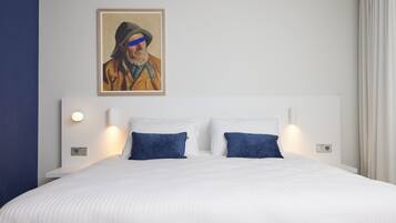 A neatly made bed with white bedding and two blue pillows, a framed painting on the wall, and bedside lamps.