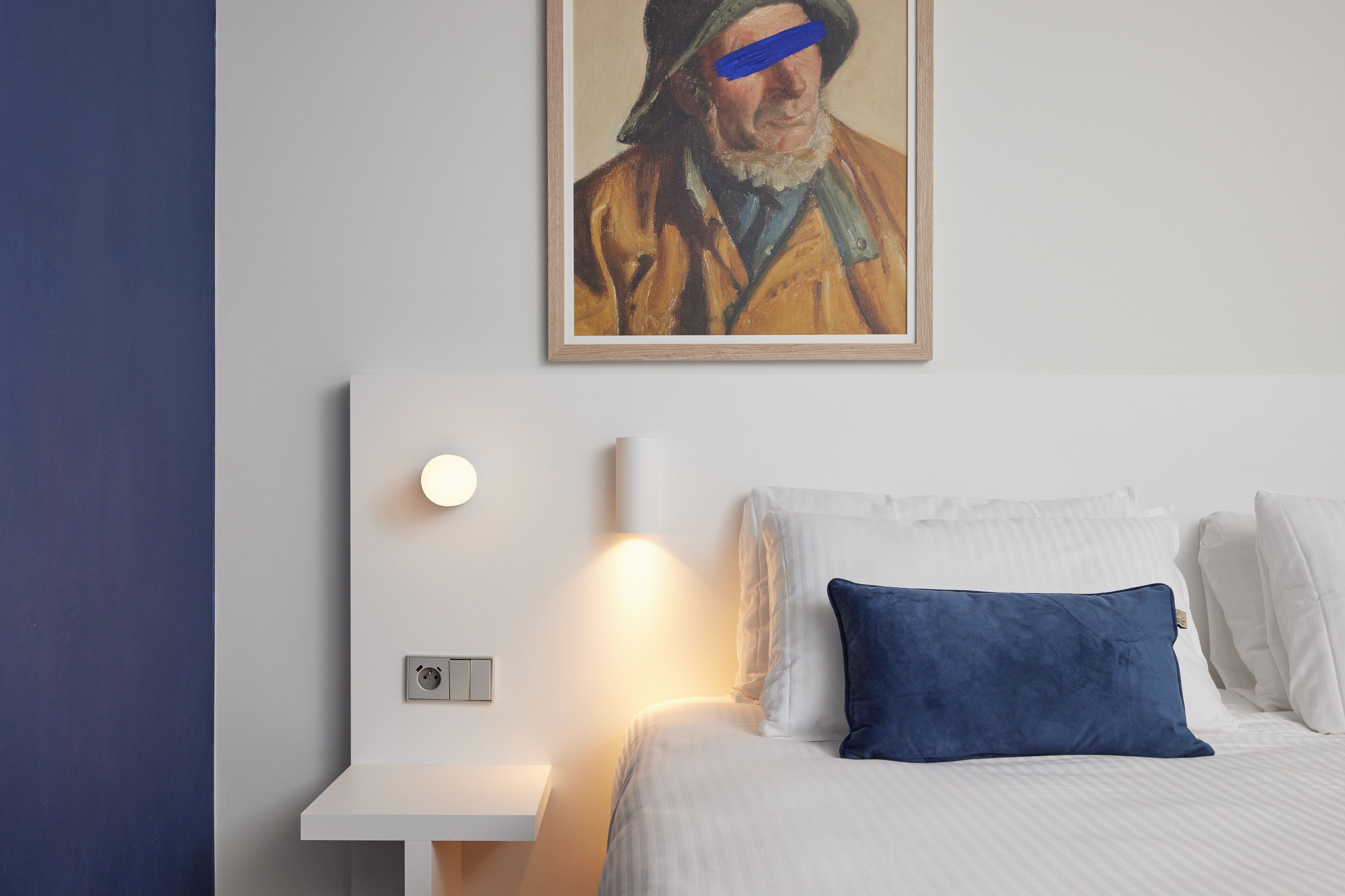 A hotel room with a bed, white pillows, a blue pillow, a painting on the wall, a bedside table, and a wall-mounted light.