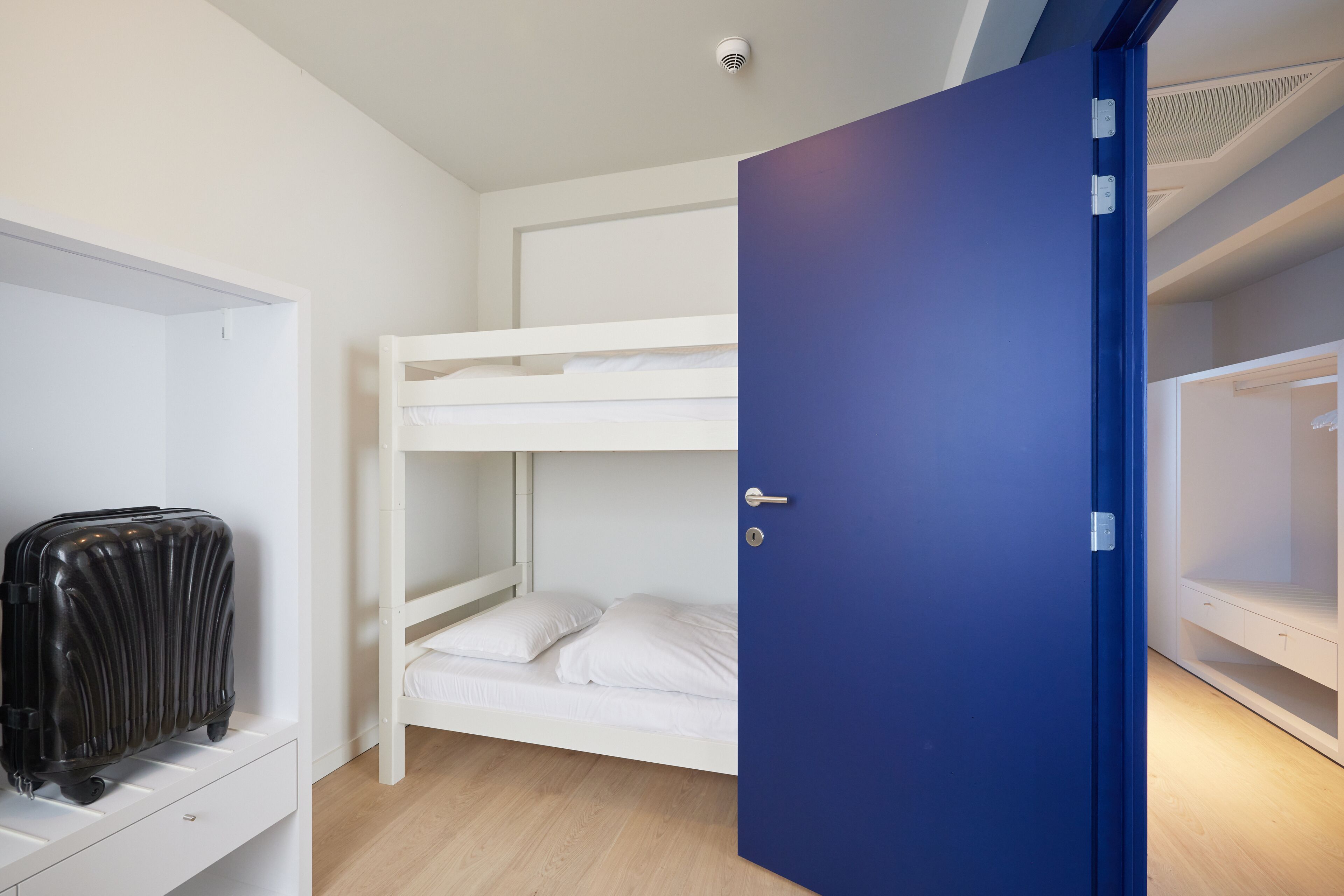 Une chambre avec un lit superposé, une porte bleue, une valise et une armoire blanche.