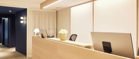 A hotel reception desk with a wooden counter, a desk lamp, a vase with flowers, a laptop, and a phone.