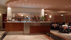 A bar area with a curved counter, multiple bottles of liquor, and a potted plant. There are tables and chairs arranged for dining in the background. - Hotel Kyriaki (Chania)