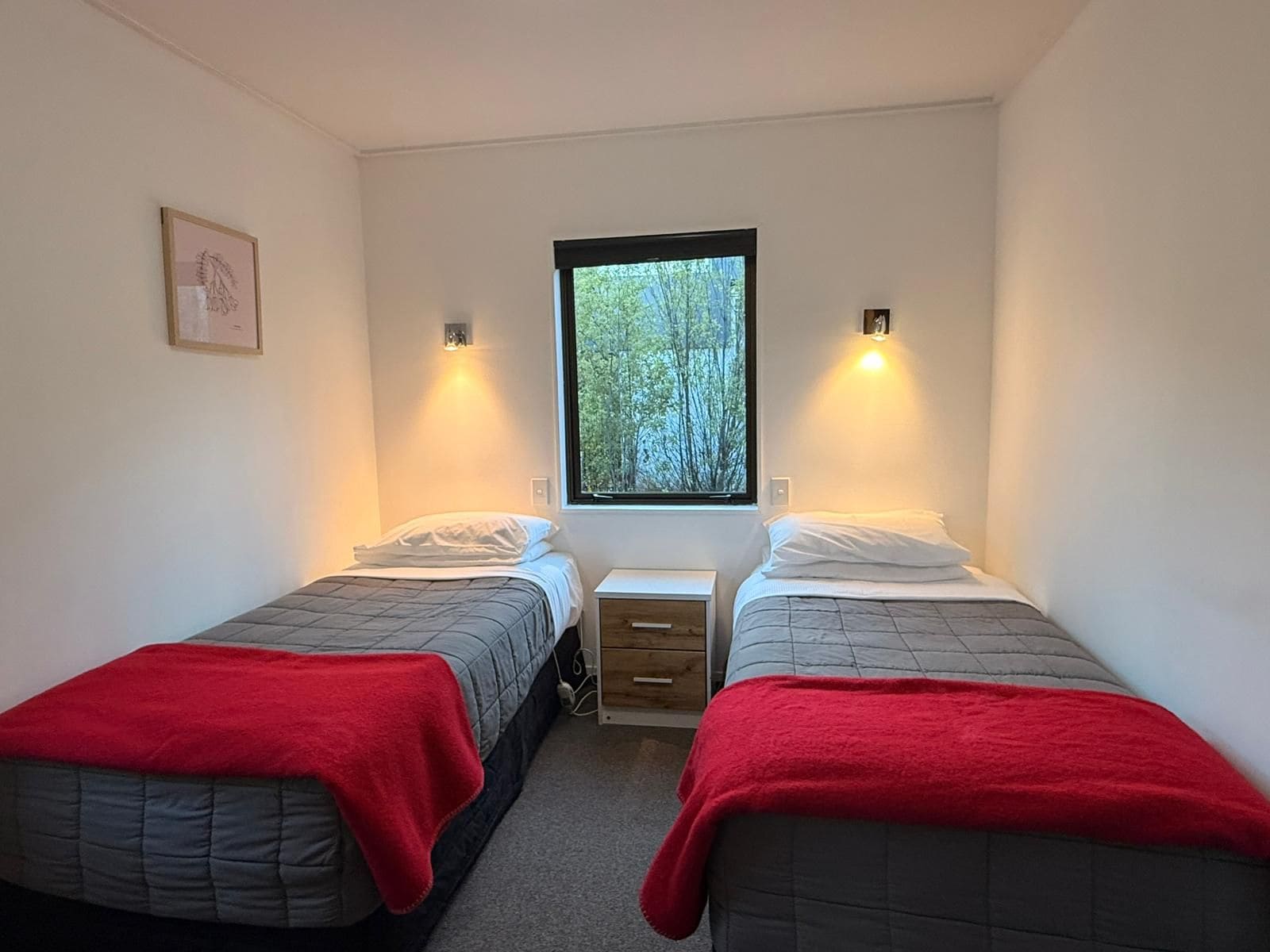 A room with two single beds, each with a red blanket, a window with a view of trees, and two wall-mounted lights.