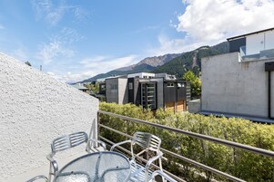 A balcony with metal chairs and a glass table, overlooking a mountainous landscape and modern buildings.