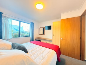 A bedroom with a bed, a TV, a wooden wardrobe, and a window with curtains. - Amity Serviced Apartments (Queenstown)