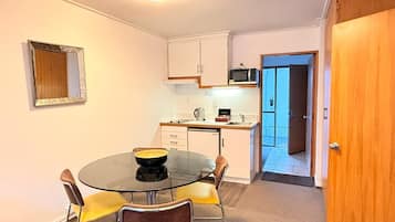 A compact kitchen with a microwave, kettle, and a small refrigerator. There is a round glass table with four chairs, one of which has a yellow cushion. A wooden door is visible on the right.