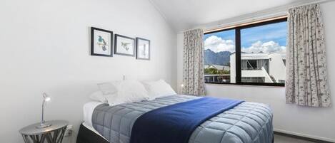 A bedroom with a bed, a bedside table, a lamp, a window with curtains, and framed pictures on the wall.