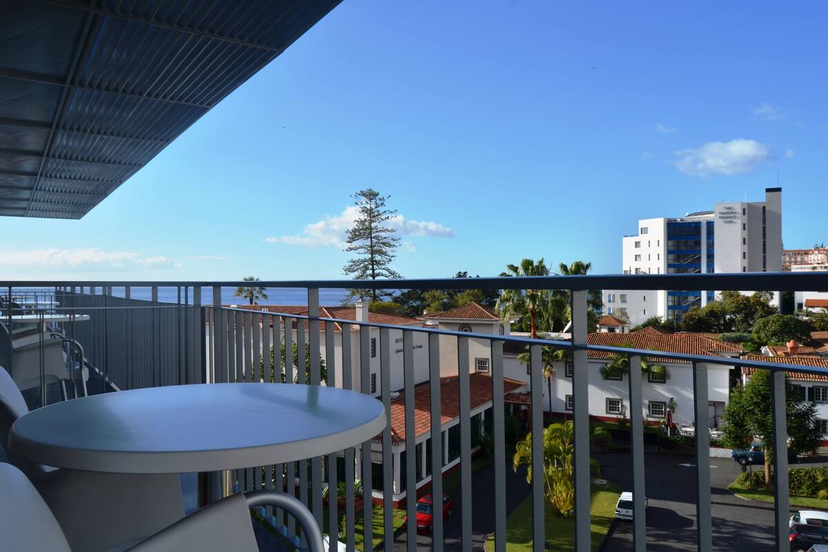 A balcony with a table and chairs, overlooking a street with buildings and a clear sky.