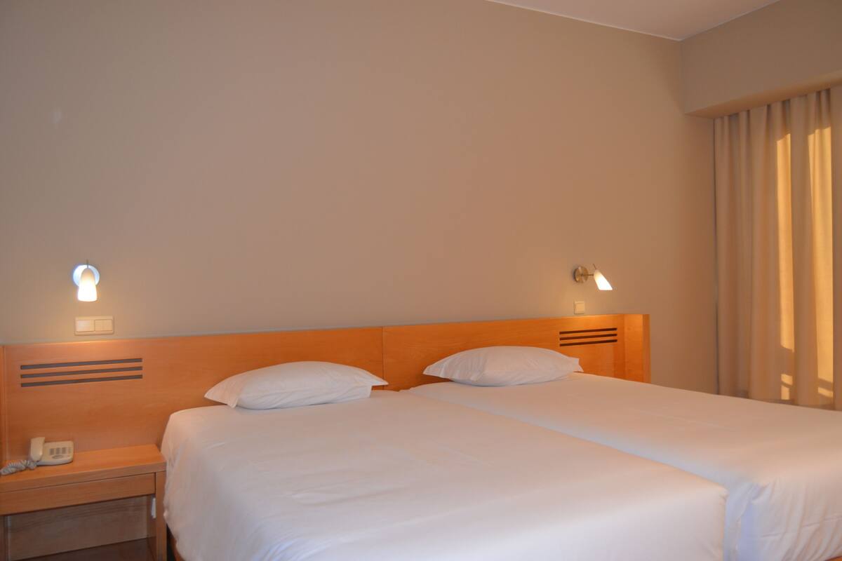 A hotel room with two beds, a wooden headboard, bedside lamps, a telephone, and a window with curtains.