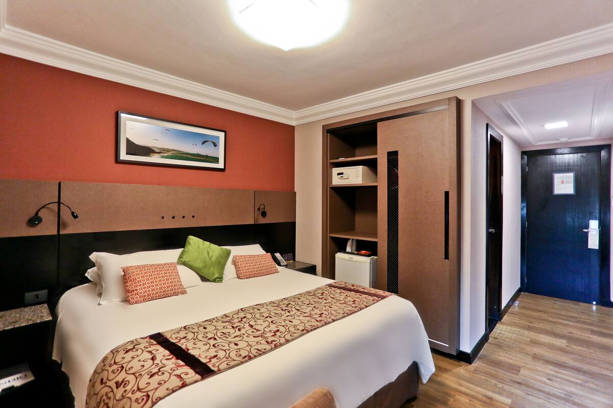 A hotel room with a large bed, a nightstand, a desk, a wardrobe, and a door leading to another room.