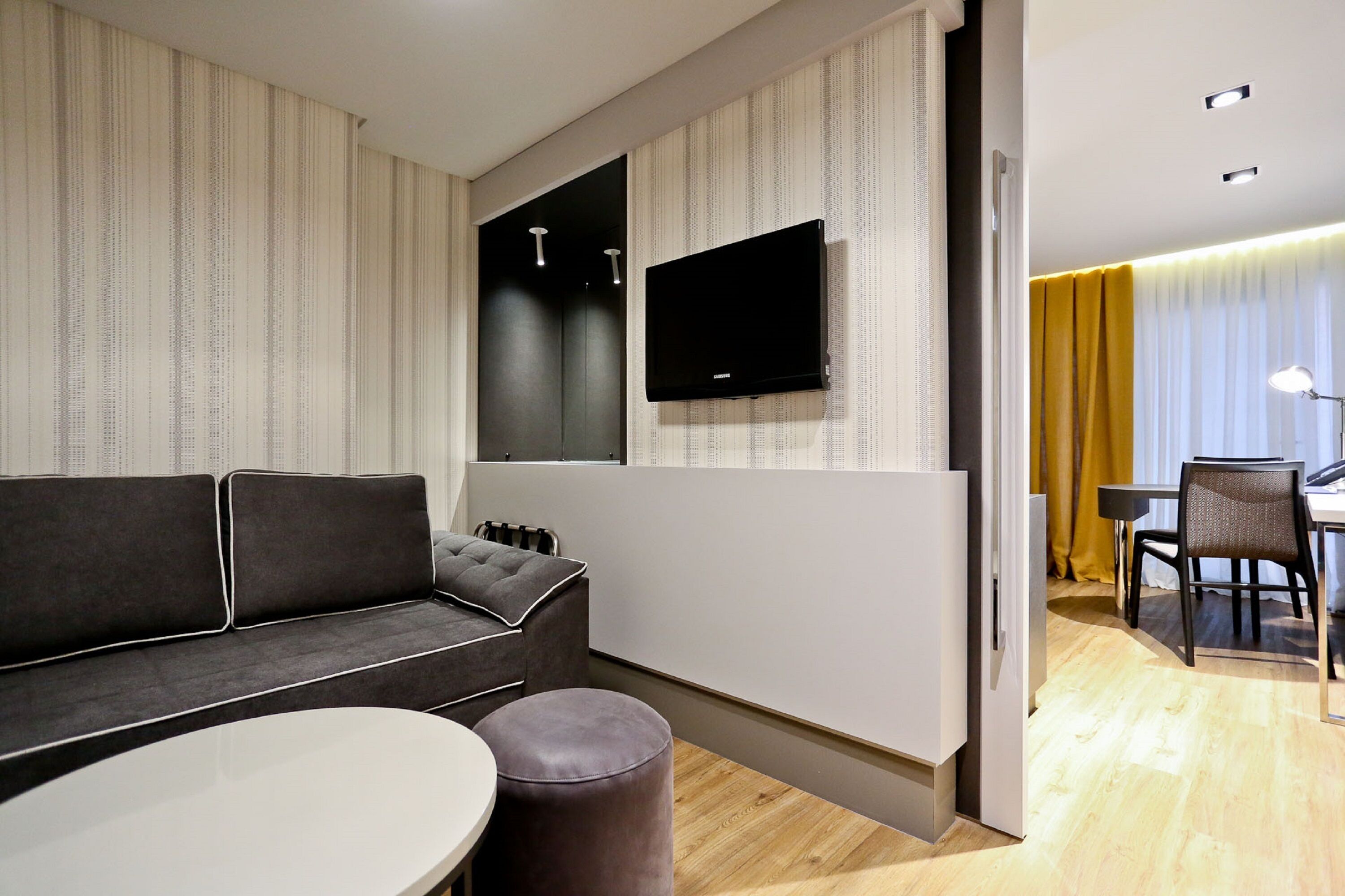 A modern hotel room with a sofa, a round table, a flat-screen TV mounted on the wall, and a dining area with a table and chairs.