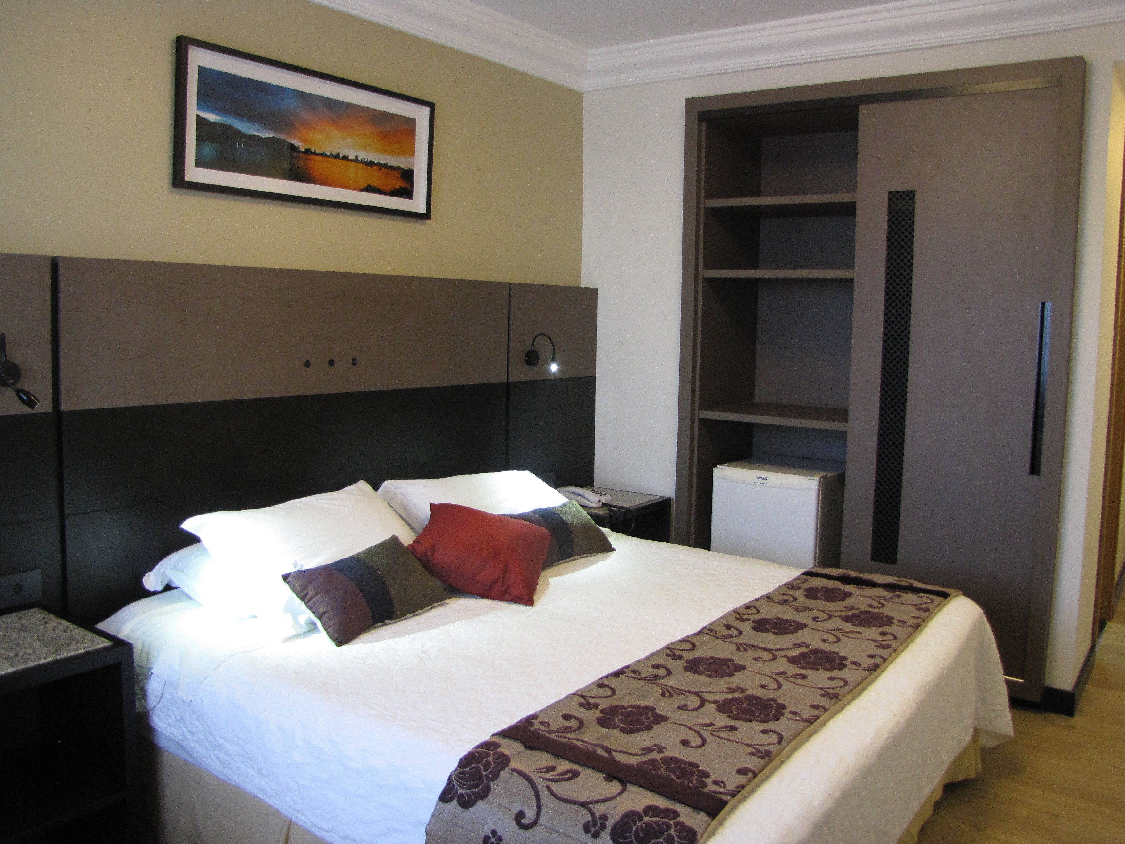 A hotel room with a bed, a nightstand, a desk, a wardrobe, and a picture on the wall.