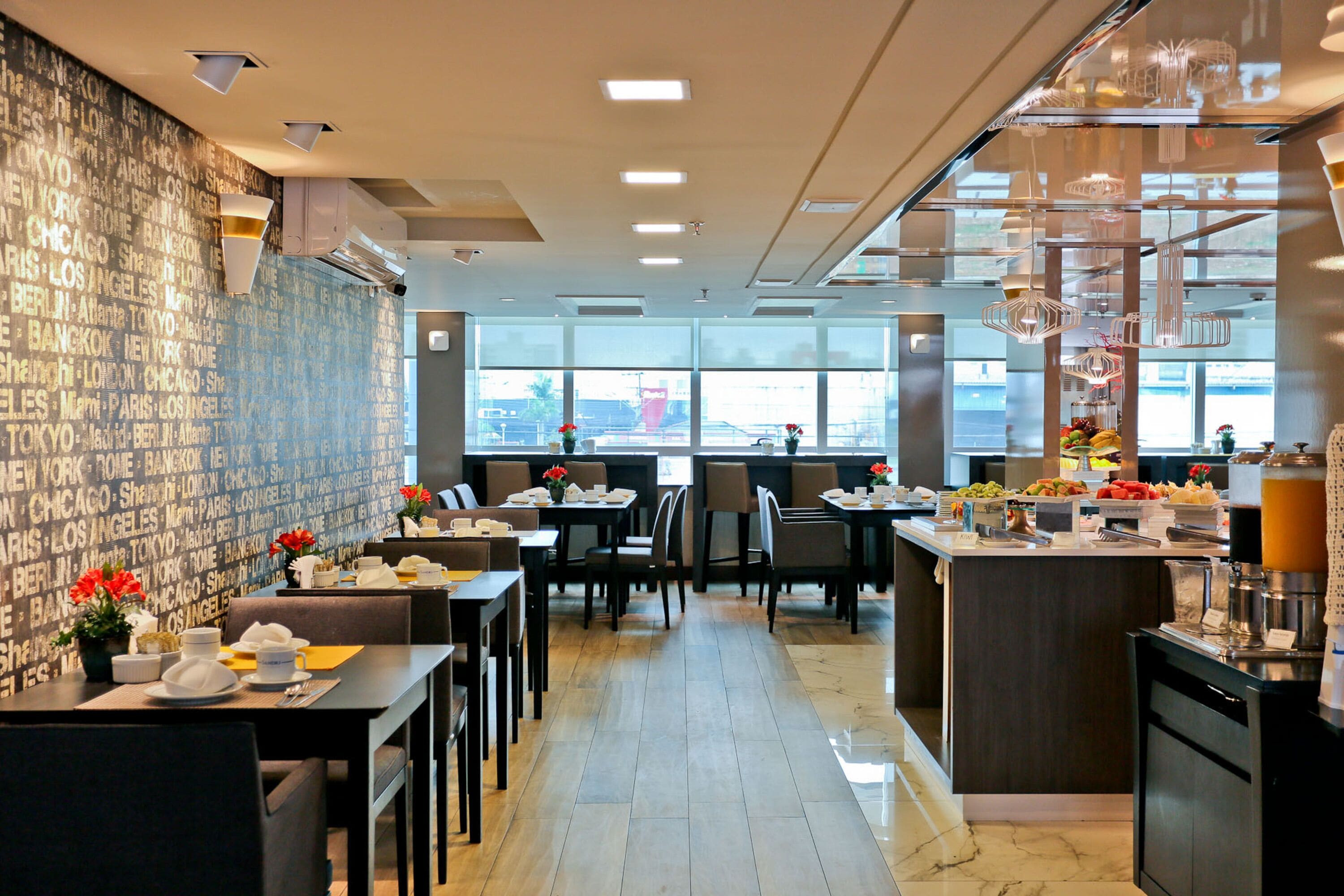 A modern restaurant with a wall of city names, dining tables with chairs, a kitchen area, and large windows.