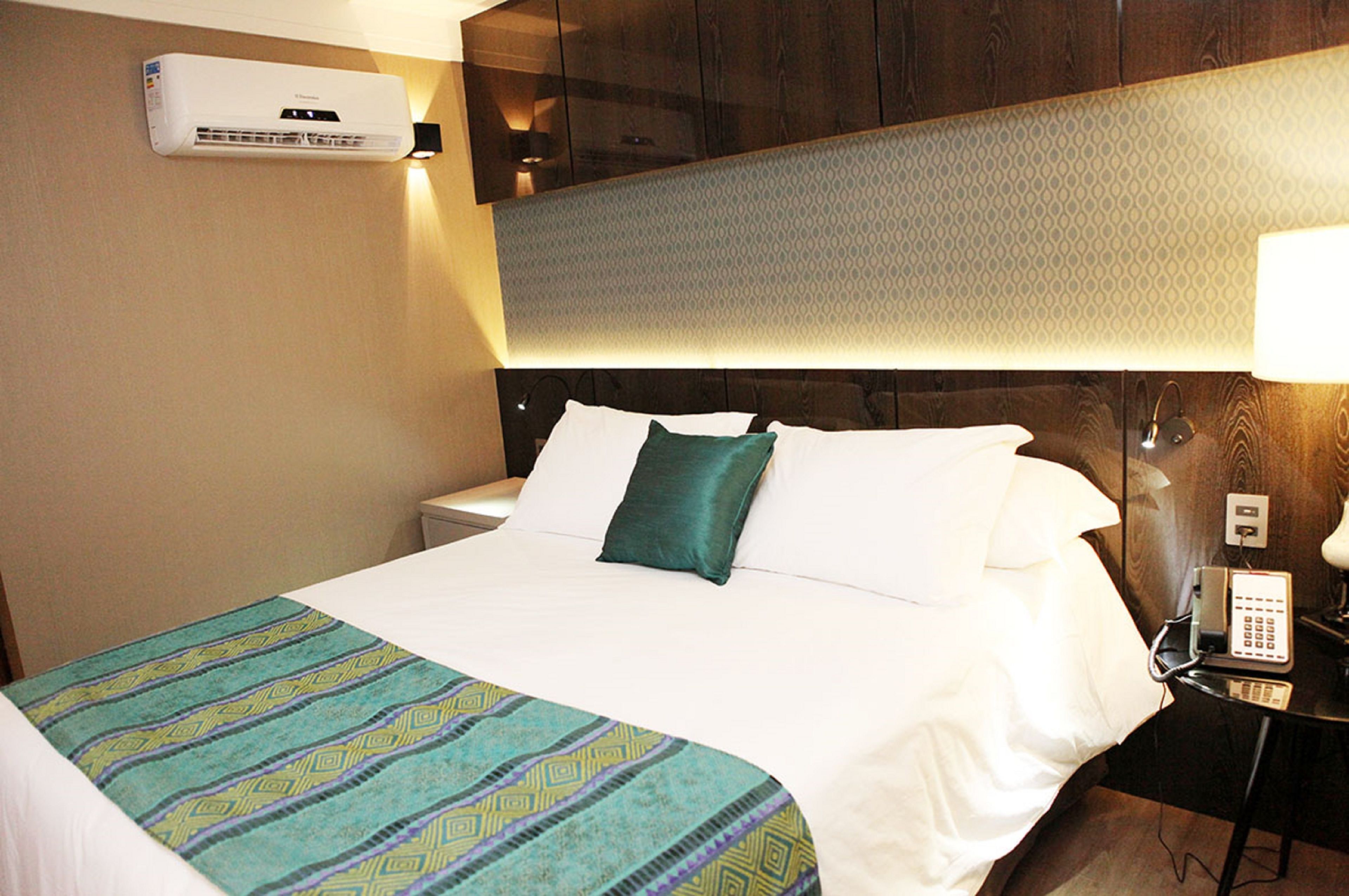 A hotel room with a bed, a bedside table with a lamp, a telephone, and an air conditioning unit on the wall.
