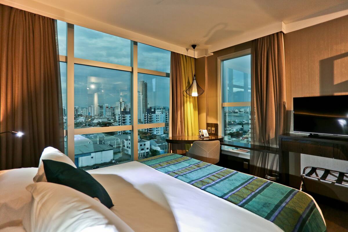 A hotel room with a large bed, a TV, a desk with a lamp, and a view of the cityscape through large windows.