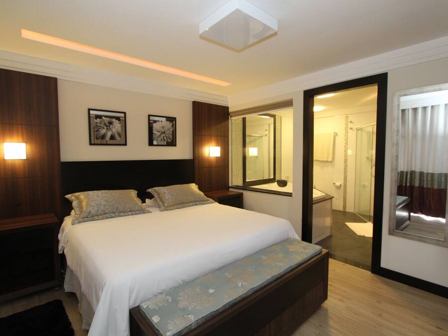 A hotel room with a large bed, bedside tables, a bench, a mirror, and a bathroom visible through an open door.