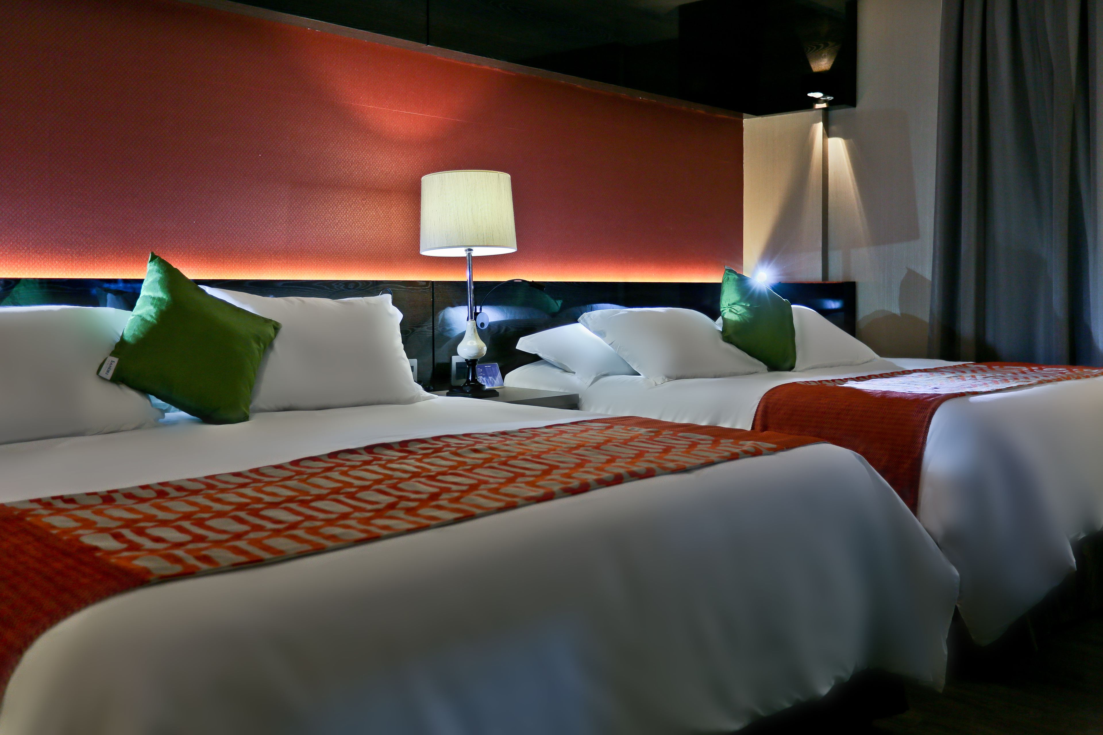 A hotel room with two beds, each with white bedding and green pillows. There is a red patterned throw blanket on one bed. A lamp is on the nightstand beside the beds.