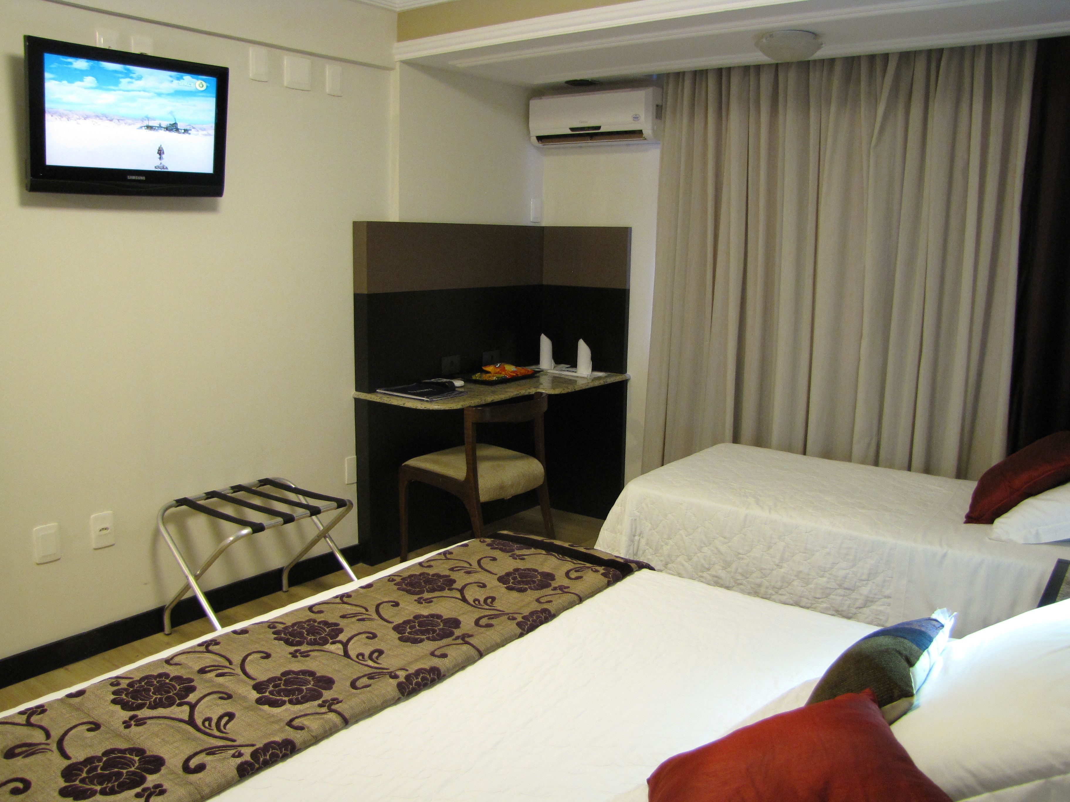 A hotel room with two beds, a desk, a chair, a TV, and a window with curtains.