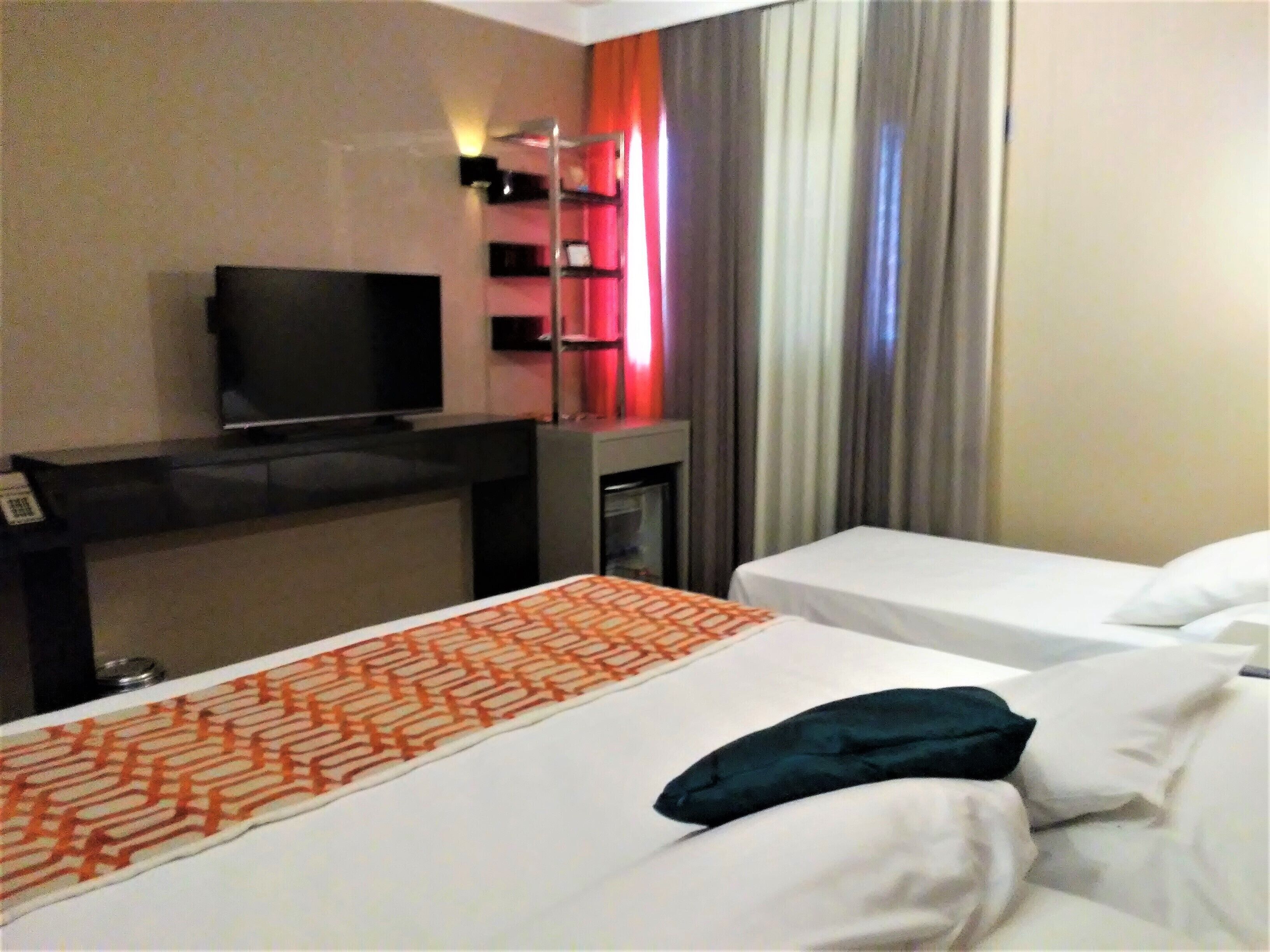 A hotel room with a bed, a TV, a desk, and a shelf with items.