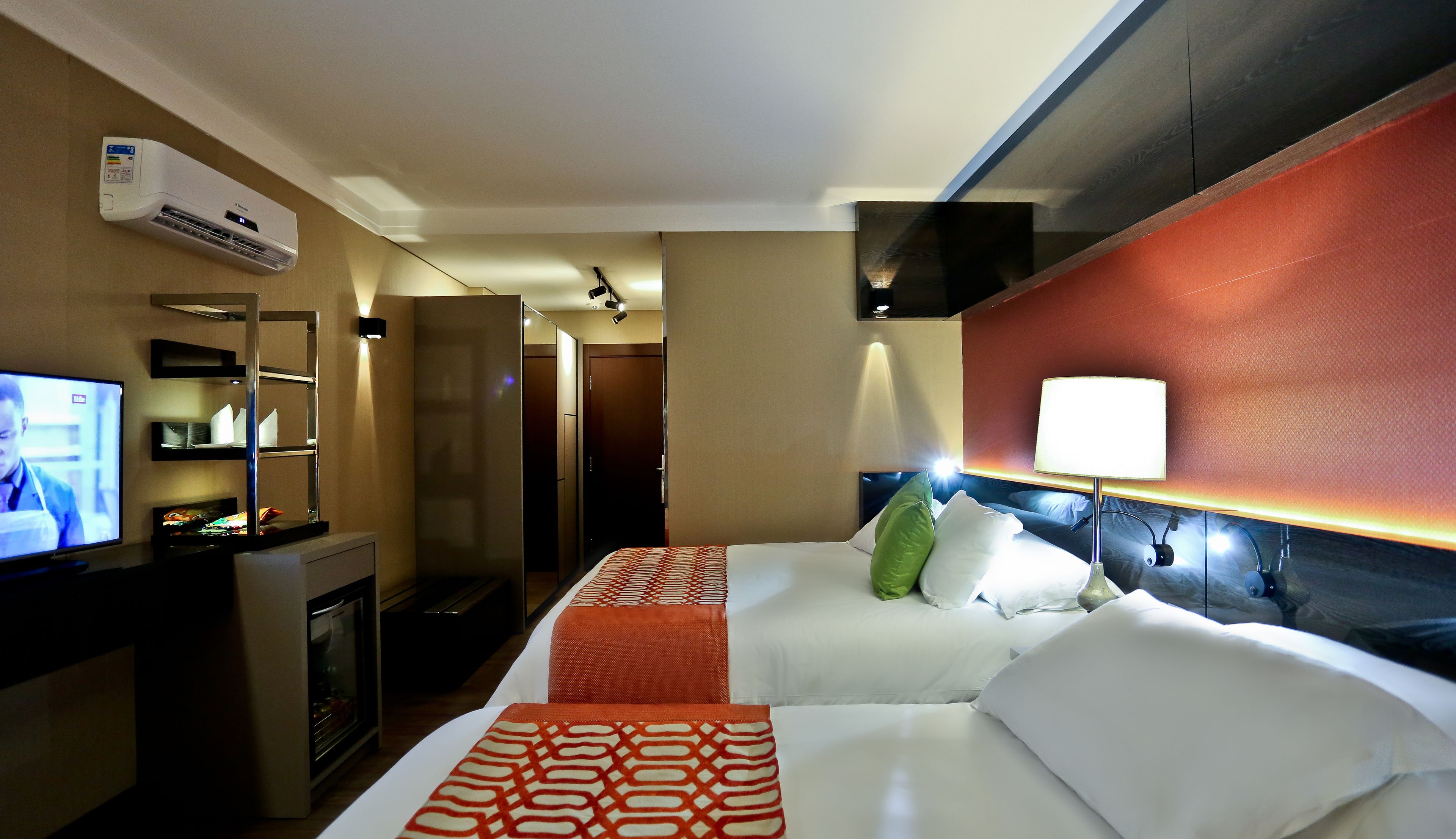 A hotel room with a bed, bedside table, TV, air conditioning unit, and a wall-mounted lamp.