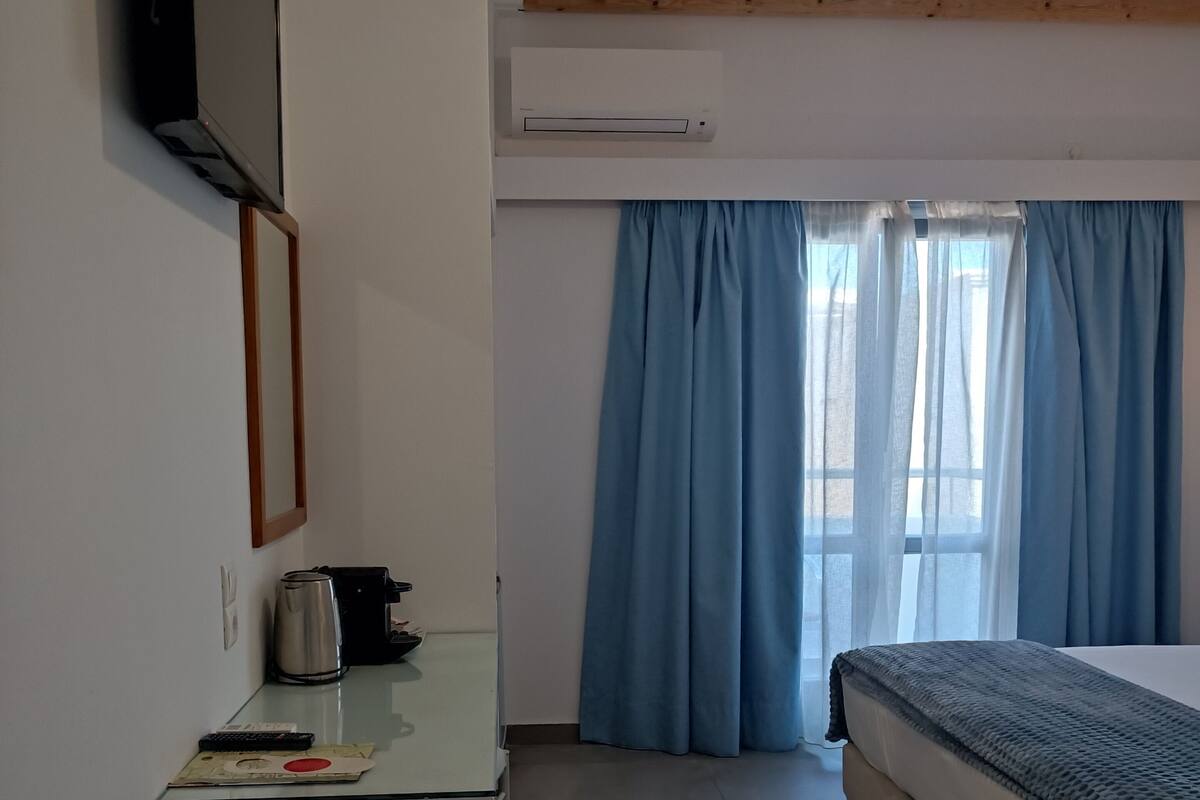 A hotel room with a bed, a small table with a kettle, a TV mounted on the wall, and a window with blue curtains.