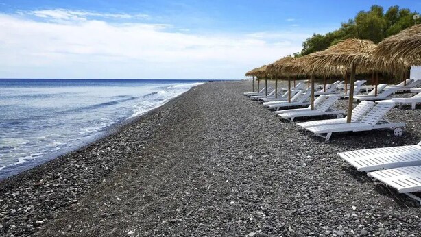 Una playa con guijarros, sillas de descanso blancas y sombrillas de paja.