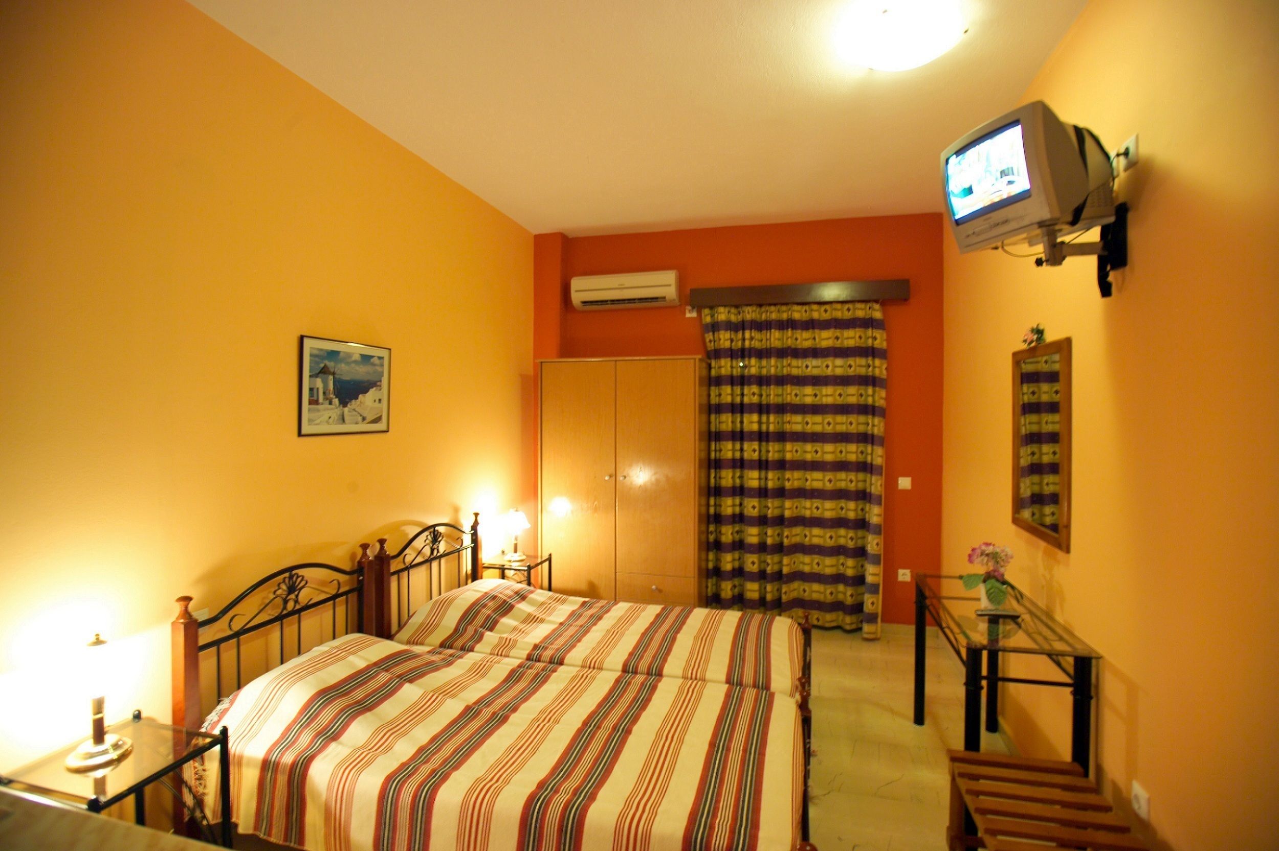 Une chambre à coucher comprenant un lit, une table de chevet, une lampe, un appareil de climatisation, un téléviseur, des rideaux et un tableau encadré accroché au mur.