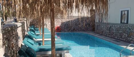 Une piscine avec des chaises longues, un mur en pierre et un parasol en chaume.