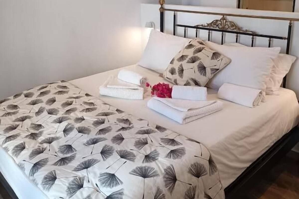 A neatly made bed with a leaf-patterned bedspread, a white headboard, and a small bedside table with a lamp.