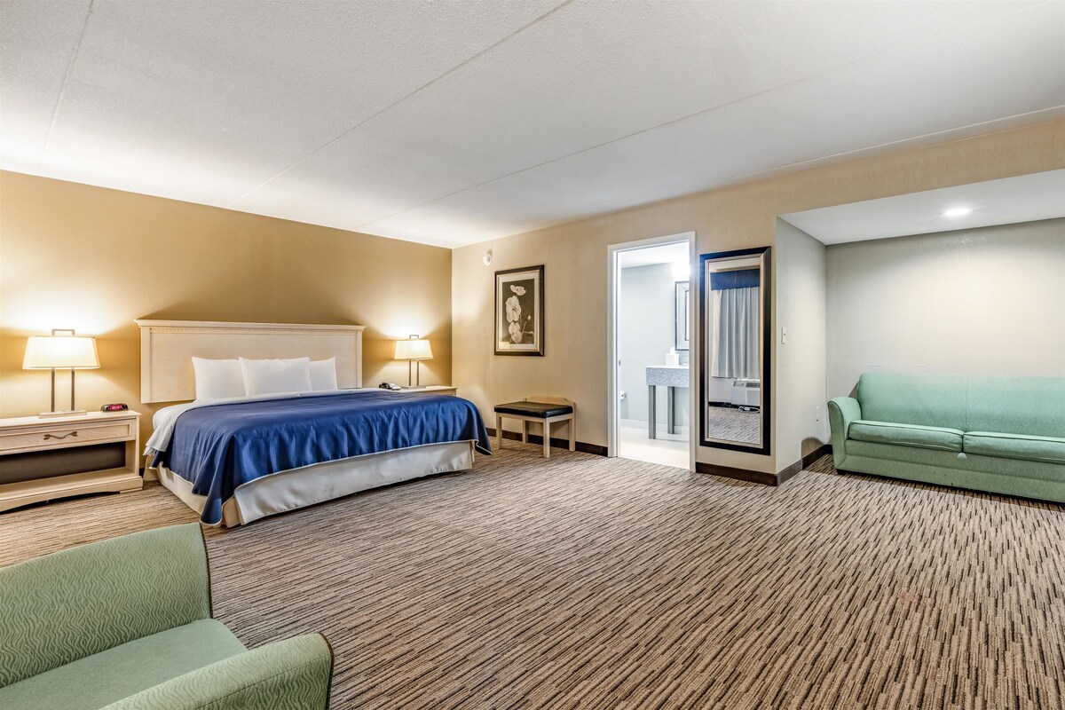 Habitación de hotel con cama, mesitas de noche, un sillón verde, sofá y una puerta que da a un balcón.