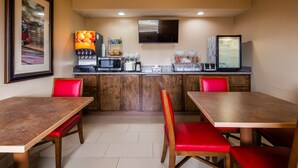 Una cocina compacta con armarios de madera, un microondas, un tostador y un dispensador de zumo. Hay dos sillas rojas y una mesa de madera.