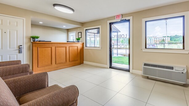 Un área de recepción con un escritorio de madera, una barra de recepción, una sala de espera con sillones tapizados en marrón, un ventanal con vistas a edificios y una puerta señalizada con “EXIT”.