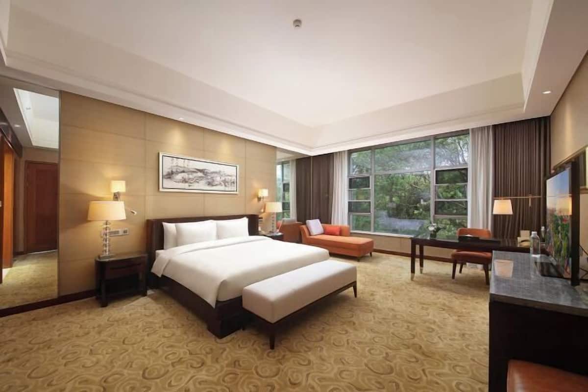 Een hotelkamer met een groot bed, een bureau, een stoel, een bank en een groot raam met uitzicht op groen.