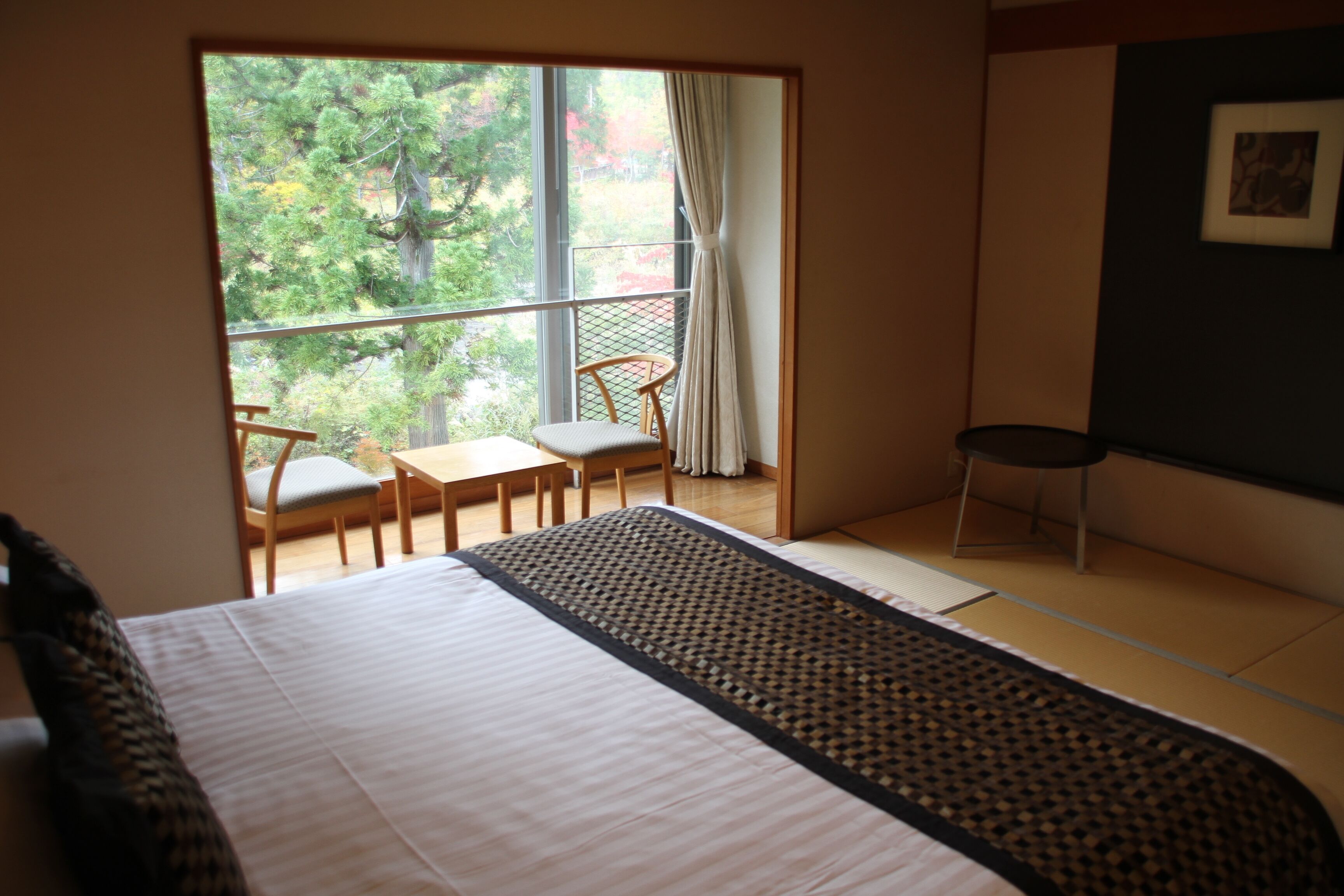 A room with a bed, a small table, two chairs, a view through a window, and a picture on the wall.